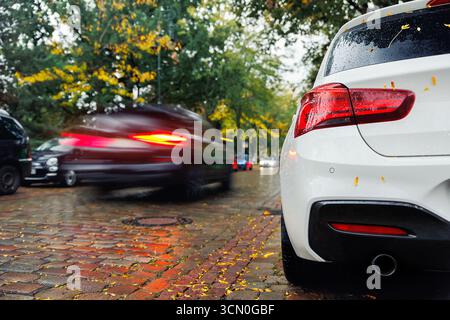 Moody Scenic feu arrière vue sur le pare-brise de voiture blanc moderne couvert de feuilles jaunes dorées tombées garées rue humide de ville automne jour pluvieux. Couvert Banque D'Images