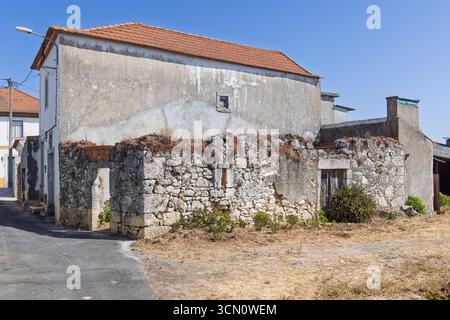 Une vue latérale d'un bâtiment portugais abandonné avec des murs de pierre en ruine, un toit de tuiles rouges et de l'herbe sèche à l'avant, montrant son état de négligence Banque D'Images