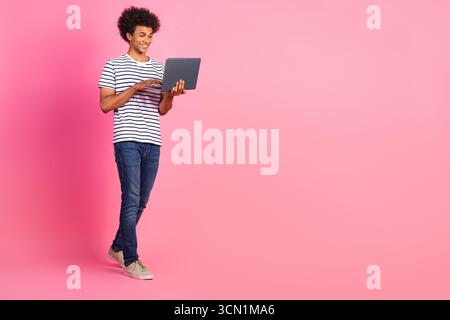 Jeune homme de race mixte vêtu de vêtements décontractés à l'aide d'un ordinateur portable, debout sur un fond rose et souriant Banque D'Images