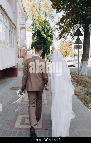 Mariés et mariés marchent main dans la main le long d'un trottoir de la ville, mariés tenant un bouquet blanc, célébrant leur jour de mariage dans un cadre urbain. Banque D'Images