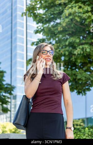 Femme d'affaires souriante et parlant sur son smartphone tout en marchant devant le bâtiment Regione Lombardia dans le quartier financier de Milan, en Italie Banque D'Images