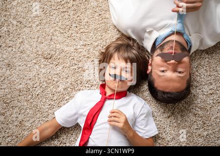 Père et fils avec moustache en papier couché sur un tapis, vue de dessus. Espace pour le texte Banque D'Images