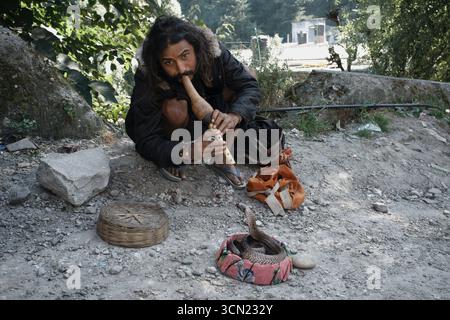 Manali, Himachal Pradesh, Inde - 11 octobre 2011 : Portrait d'un charmeur de serpent indien regardant la caméra tout en étant assis sur le hantail et en jouant au pungi. Cobra assis dans un petit panier pour la performance de rue Banque D'Images