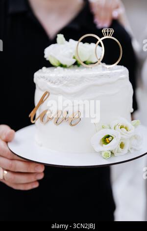 Élégant gâteau de mariage avec des anneaux et des fleurs blanches, tenu par les jeunes mariés dans la célébration Banque D'Images
