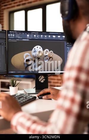 Ingénieur masculin noir travaillant sur le prototype de machines de construction en utilisant un logiciel de CAO dans le home studio moderne. Le moniteur de bureau affiche l'interface technique et les outils de modélisation de précision. Banque D'Images