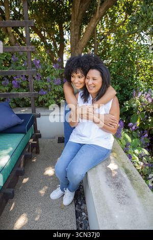 Embrasser la mère et la fille enveloppant autour des épaules sur le patio par treillis avec des fleurs violettes Banque D'Images