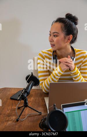Femme asiatique dans la chemise rayée assis à table dans le studio en utilisant des micros et des écouteurs pour ordinateur portable Banque D'Images