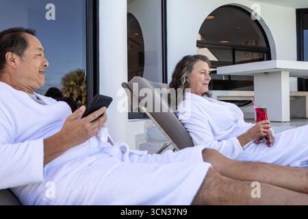 Couple diversifié s'inclinant sur des chaises longues à la terrasse du spa tenant smartphone et boisson dans le verre Banque D'Images