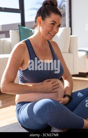 Femme enceinte assise les jambes croisées sur un tapis de yoga dans le salon, berçant le ventre et souriant Banque D'Images