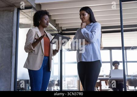 Divers collègues discutant des détails du projet au bureau près de la cloison en verre avec tablette et ordinateur portable Banque D'Images