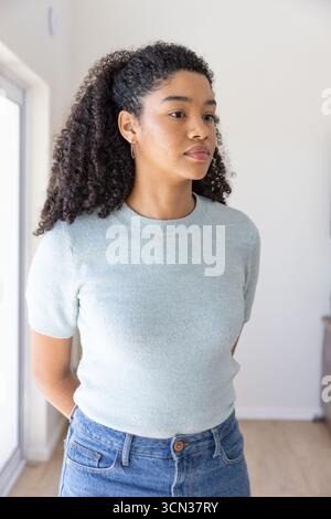 Femme afro-américaine debout dans la maison ensoleillée portant un pull bleu, un Jean à côté du capteur de fenêtre Banque D'Images