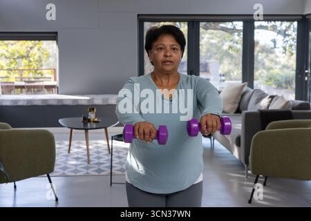 Femme indienne senior soulevant des haltères violets dans le salon par une table basse ronde Banque D'Images