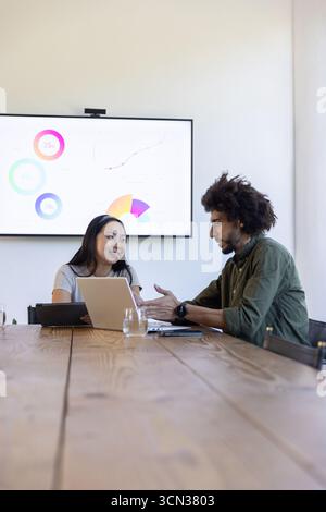 Divers collègues dans la salle de conférence discutent des données affichées, examinent l'ordinateur portable à table Banque D'Images
