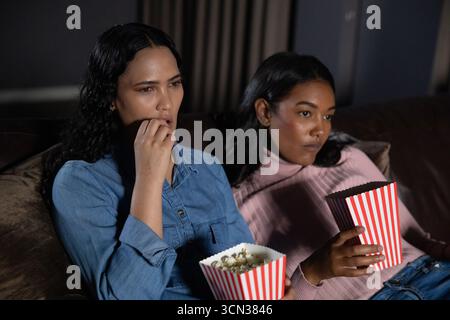 Diverses amies féminines assis sur un canapé en cuir dans le salon mangeant du pop-corn à partir de boîtes rayées Banque D'Images