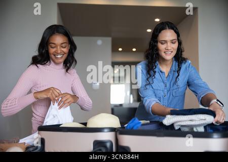 Diverses amies féminines emballant des valises sur le comptoir de cuisine, organisant des vêtements, des chapeaux de paille Banque D'Images