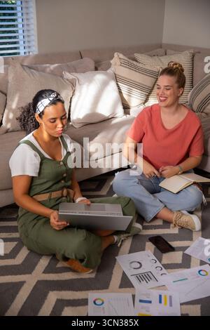 Amies féminines collaborant sur le projet sur le tapis géométrique dans le salon avec des graphiques d'ordinateur portable Banque D'Images