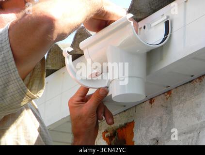 Entrepreneur installant un support de gouttière de toit en plastique pour le tuyau d'évacuation du bec verseur. Gouttière de toit en plastique, gouttière de pluie et drainage par Handyman Hands. Banque D'Images