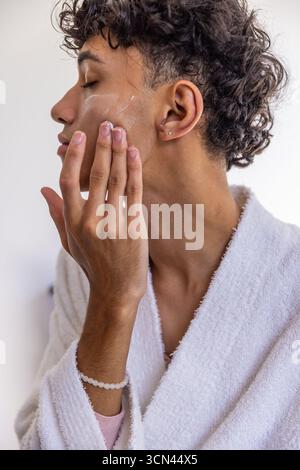Homme en peignoir blanc appliquant la crème à la joue dans la salle de bain portant bracelet et clous d'oreille Banque D'Images