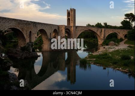 Par une chaude soirée de juin, la paisible rivière Fluvià reflète les arches inégales et la tour de défense centrale du Viejo médiéval reconstruit, Pont Vell ou Vieux Pont, construit sur les rochers du lit de la rivière à Besalú, Gérone, Catalogne, Espagne. La structure fortifiée inclinée du 11ème siècle ce, d'environ 105m ou 344 pieds de long, est un monument emblématique catalan et un emblème populaire de la ville antique. Banque D'Images