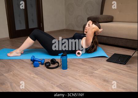 Femme sur tapis de yoga à la maison effectuant des exercices avec rouleau en mousse pour un dos sain, suite à la formation de fitness en ligne sur ordinateur portable, bien-être et LIF actif Banque D'Images