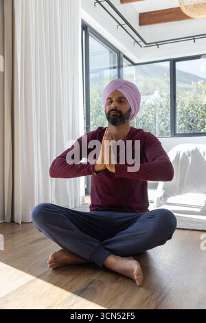 Homme indien adulte moyen assis sur plancher en bois dans la chambre à côté de rideaux blancs, porte coulissante en verre Banque D'Images