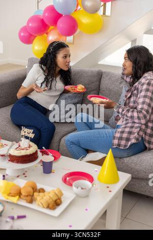 Maman diversifiée et fille adolescente célébrant sur le canapé gris dans le salon avec gâteau d'anniversaire Banque D'Images
