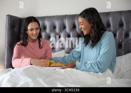 Asiatique avec des femmes afro-américaines dans des vêtements de détente partageant le plateau de petit déjeuner dans le lit avec du jus d'orange Banque D'Images