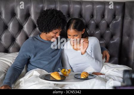 Couple diversifié appuyé contre la tête de lit sur le lit tout en partageant plateau, croissants et fruits Banque D'Images