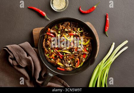 Steak de boeuf frit finement tranché avec des poivrons dans une poêle en fonte sur fond brun foncé. Vue de dessus, pose à plat Banque D'Images