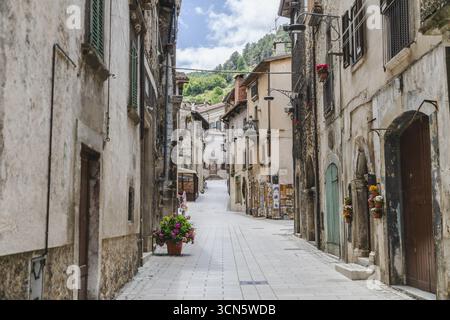 Vue des bâtiments en pierre vieillie bordent une rue étroite avec des pots de fleurs, sous un ciel lumineux, menant à des collines verdoyantes lointaines, Scanno, Abruzzes, Italie. Banque D'Images