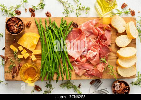 Une planche de charcuterie magnifiquement agencée avec des tranches de jambon, des blocs de fromage, des asperges fraîches et des noix croquantes. Parfait pour déguster avec frien Banque D'Images