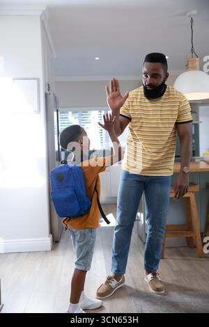 Père et fils afro-américains en haut-fiving dans la zone d'entrée de la cuisine à la maison avec sac à dos d'école bleu Banque D'Images