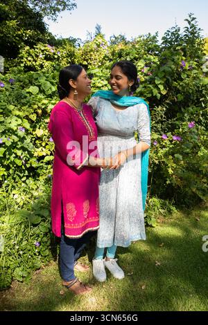 Mère indienne et fille debout, se tenant la main et souriant sur la pelouse du jardin sous la lumière du soleil Banque D'Images