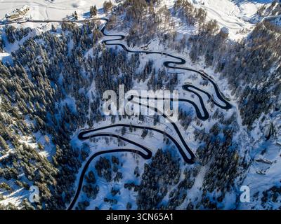 Vue aérienne d'une route sinueuse serpente à travers une forêt enneigée, un contraste frappant avec le paysage blanc lumineux, col de Maloja, Maloja, Suisse. Banque D'Images