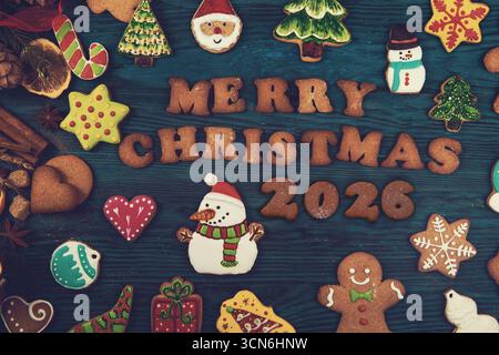 Des biscuits de Noël colorés en forme de bonhommes de neige, d'arbres, d'étoiles et de pain d'épice entourent le texte Joyeux Noël 2026 sur un fond en bois. Banque D'Images