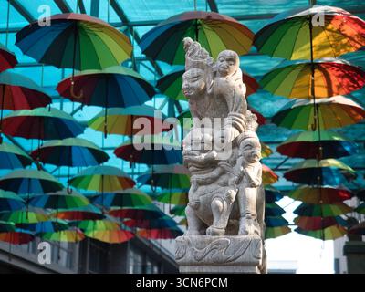 Statue chinoise traditionnelle avec des parapluies colorés - Une statue en pierre détaillée d'une figure chinoise traditionnelle se tient sous un plafond de couleur vibrante Banque D'Images