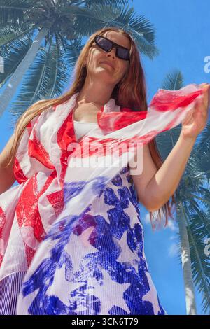 Une femme portant des lunettes de soleil et une écharpe de style drapeau américain pose sous de grands palmiers par une journée ensoleillée. Fond tropical, dynamique, patriotisme. Banque D'Images