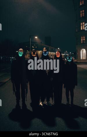 Un groupe d’individus portant des masques lumineux debout sur une rue faiblement éclairée la nuit, évoquant une atmosphère mystérieuse et énigmatique avec un fès Banque D'Images