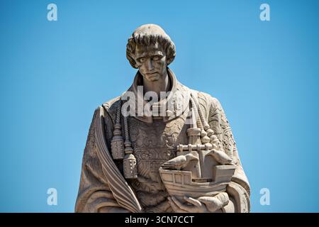 Statue de Saint Vincent Holding Ship Lisbonne Portugal // LISBONNE, Portugal — la statue de Saint Vincent (São Vicente) est sculptée dans du calcaire clair et représente le saint patron de Lisbonne tenant un modèle de navire de style carrack. Ce navire symbolise les vaisseaux qui, selon la légende, transportèrent ses reliques à la ville au XIIe siècle. La statue, située au point de vue Miradouro das Portas do sol dans le quartier historique d'Alfama, montre le saint dans des robes liturgiques élaborées, faisant référence à son rôle de diacre et de martyr. En arrière-plan, les tours jumelles de l'église et du monastère de São Vic Banque D'Images