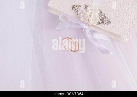 Deux anneaux de mariage en or se trouvent parmi les plis doux du voile blanc de la mariée à côté d'une belle enveloppe d'invitation de mariage. vue de dessus. Un espace de copie. Banque D'Images