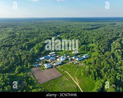 Une vue aérienne large d'un complexe agricole avec des bâtiments et des champs, situé à côté d'une vaste forêt verte et d'une rivière sinueuse sous un ciel bleu clair. Banque D'Images