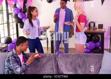 Divers amis adolescents tenant des assiettes à gâteau et des tasses sur un canapé gris dans le salon, des ballons Banque D'Images