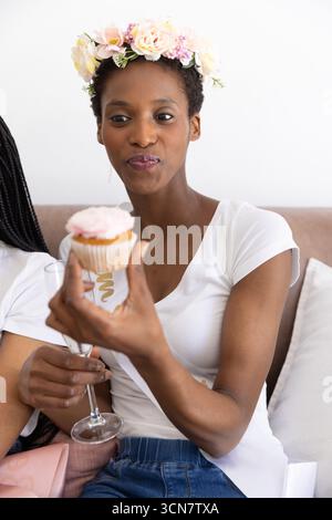 Amies afro-américaines assises sur le canapé portant une couronne, tenant un cupcake et une flûte à champagne Banque D'Images