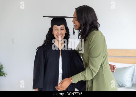 Maman diversifiée et fille adolescente debout dans la chambre portant une robe noire et un chapeau mortarboard Banque D'Images