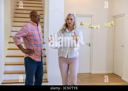 Couple senior diversifié parlant, geste dans l'entrée avec escalier en bois, bannière de bienvenue à la maison Banque D'Images