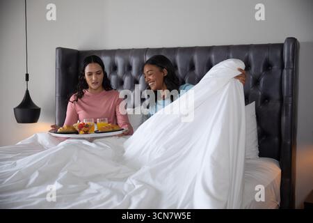 Diverses amies féminines assises sur le lit sous housse de couette avec plateau avec croissants et jus Banque D'Images