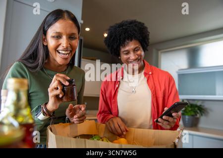 Couple diversifié déballer la boîte d'épicerie au comptoir de la cuisine, tenant le pot d'épices, vérifiant le smartphone Banque D'Images