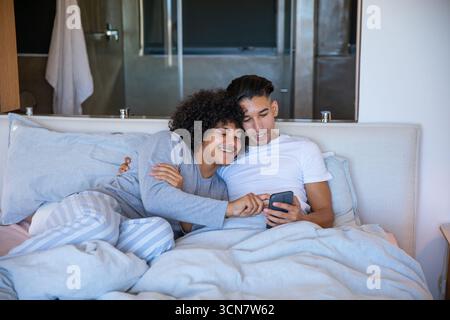 Divers partenaires masculins s'inclinant sur le lit dans une chambre moderne portant un pyjama et tenant un smartphone Banque D'Images