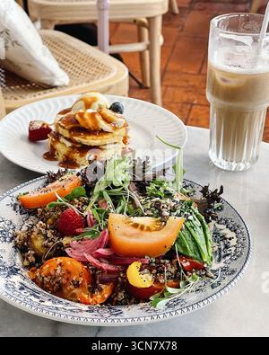 terminez la mise en place de la table du brunch. Au premier plan, une grande assiette détaillée contient une salade de quinoa saine et colorée, des crêpes et un latte Banque D'Images