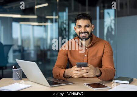 Homme d'affaires indien souriant assis à un bureau, tenant un smartphone tout en travaillant avec un ordinateur portable et une tablette, dépeignant la connectivité et le professionnalisme Banque D'Images